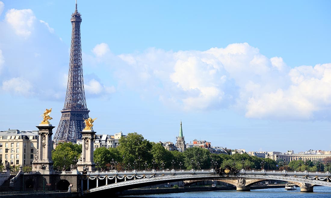 The Eiffel Tower stands tall next to a bridge and trees along the river.